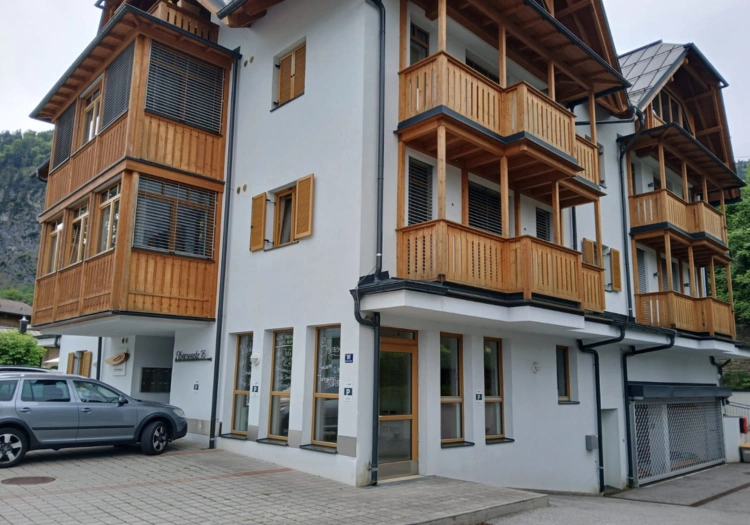 Der Weinladen befindet sich in einem modernen Gebäude mit mehreren Stockwerken und Holzverkleidungen. Das Gebäude hat weiße Wände und mehrere Balkone mit hölzernen Geländern. Direkt vor dem Gebäude befindet sich ein gepflasterter Parkplatz. Der Eingang ist verglast und neben einer Reihe von großen Fenstern im Erdgeschoss gelegen. Der Zugang zum Gebäude ist barrierefrei und auch für Rollstuhlfahrer geeignet.